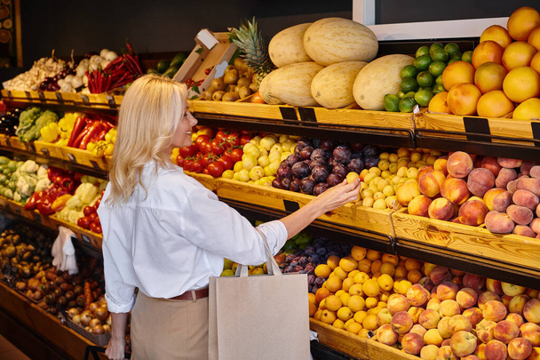 iloinen kypsä iloinen nainen jokapäiväisissä vaatteissa ostoskassilla valitsemalla hedelmiä ruokakaupassa - Valokuva, kuva