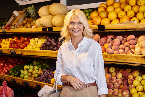 alegre mujer madura con bolsa de compras sonriendo felizmente a la cámara con tienda de comestibles en el fondo - Foto, Imagen