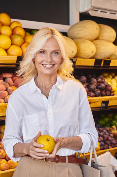 vertical shot of happy woman posing with orange and smiling at camera with grocery store on backdrop - Photo, Image