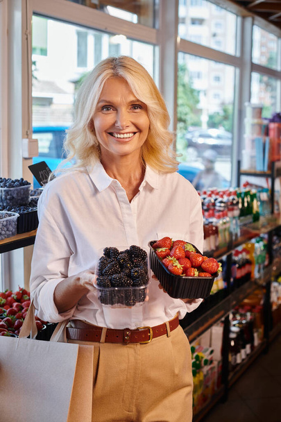 mujer madura en traje casual posando con fresas y moras en las manos sonriendo a la cámara - Foto, Imagen