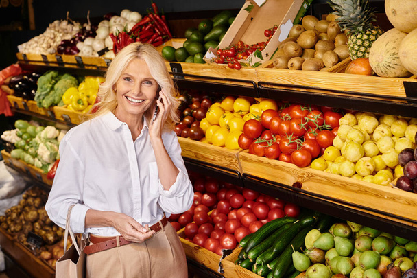 радісна приваблива жінка розмовляє по телефону в продуктовому магазині і весело посміхається на камеру - Фото, зображення