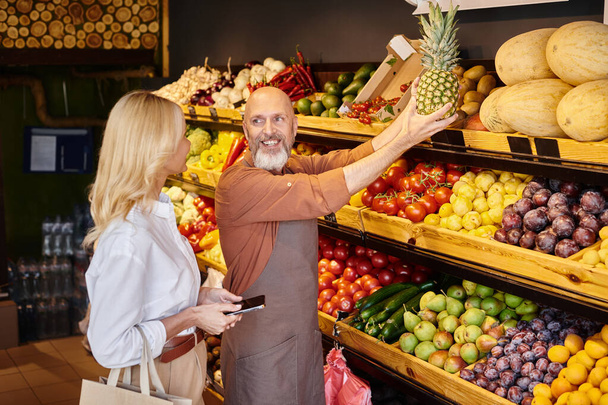 Neşeli sakallı satıcı marketin yanındaki bayan müşterisine kocaman ananas gösteriyor. - Fotoğraf, Görsel