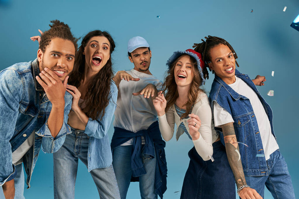 fröhliche multiethnische Freunde in trendiger Freizeitkleidung lächeln unter funkelndem Konfettiregen auf Blau - Foto, Bild