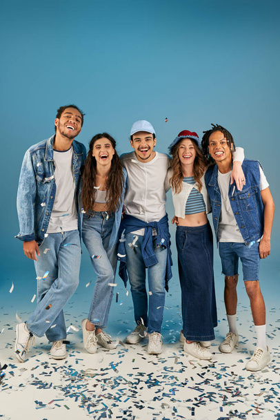 modische multiethnische Freunde in Trends und Jeans tragen lachend neben festlichem Konfetti auf blauem Grund - Foto, Bild