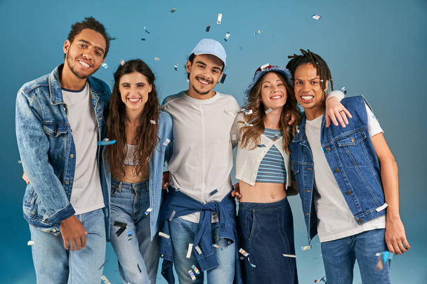 happy multiethnic friends in trendy casual outfits smiling at camera under confetti rain on blue - Photo, Image