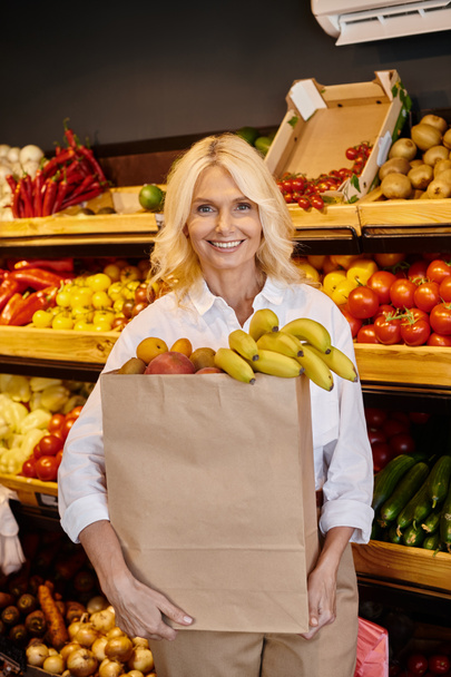 mujer madura de buen aspecto en traje casual sosteniendo bolsa de compras llena de frutas y sonriendo felizmente - Foto, Imagen