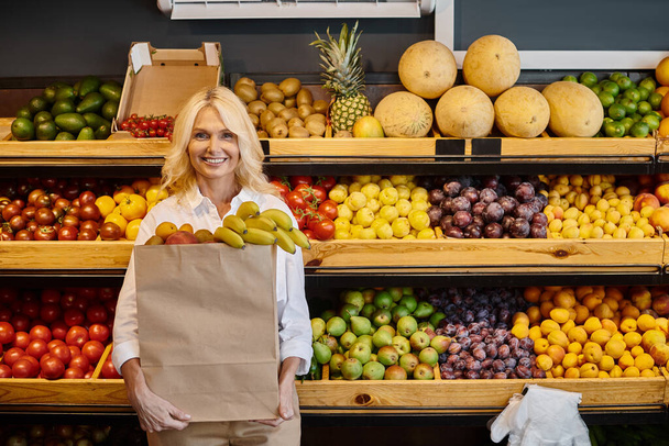 joyful attractive mature woman in everyday clothes smiling and holding shopping bag with bananas - Photo, Image