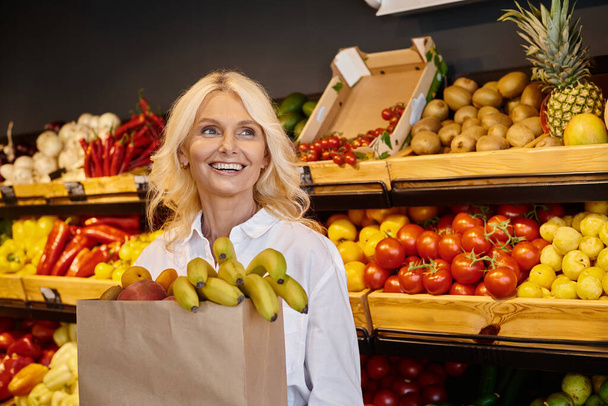 fröhliche Seniorin posiert mit Einkaufstasche voller frischer Früchte und schaut weg - Foto, Bild