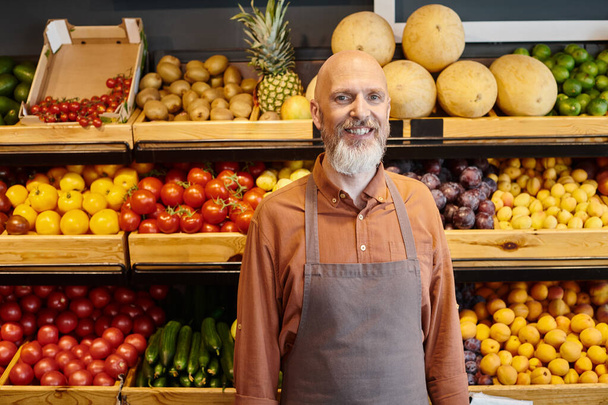 veselý vousatý zralý prodejce šťastně se usmívá na kameru a pózuje s stánkem s potravinami na pozadí - Fotografie, Obrázek