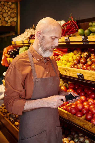 vendeur barbu mature regardant les étiquettes de prix dans ses mains avec étal d'épicerie avec des fruits sur fond - Photo, image