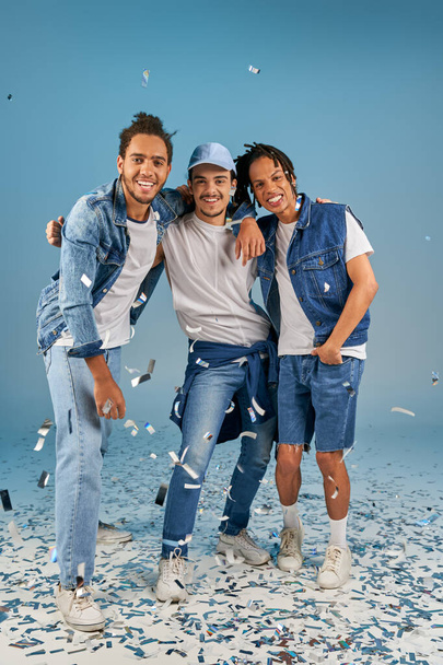 vreugdevolle interraciale vrienden in stijlvolle denim kleding omarmen onder glanzende confetti regen op blauw - Foto, afbeelding
