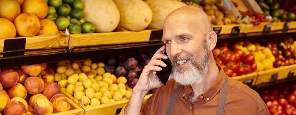 gray bearded mature salesman talking cheerfully by phone with fruits and vegetables on background - Photo, Image