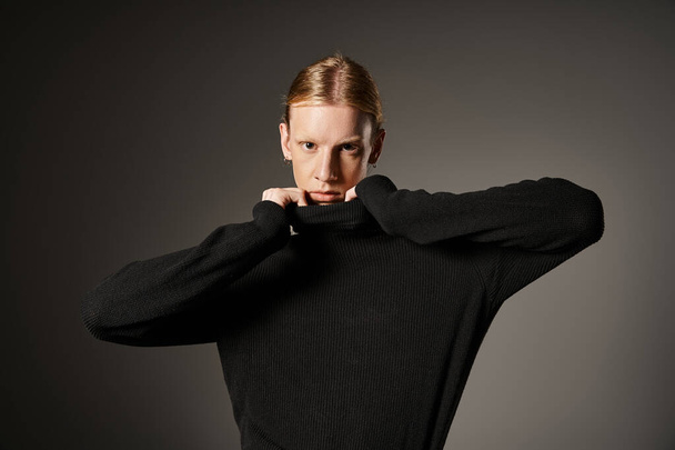 attractive young non binary person touching collar of black turtleneck and looking at camera - Photo, Image