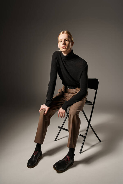 atractivo joven modelo no binario en cuello alto negro sentado en silla negra y mirando a la cámara - Foto, Imagen