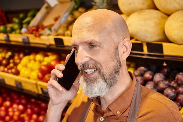 wesoły dojrzały sprzedawca rozmawiający aktywnie przez telefon z zamazanym stoiskiem spożywczym na tle - Zdjęcie, obraz