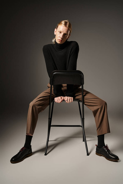 houkutteleva ei-binary henkilö b puuttuu poolopaita ja ruskea housut istuu musta tuoli, muoti - Valokuva, kuva