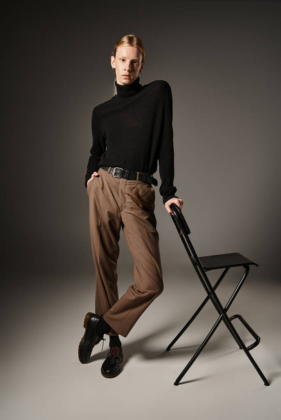 junge nicht binäre Person mit Pferdeschwanz posiert neben schwarzem Stuhl mit der Hand in der Tasche, Mode - Foto, Bild