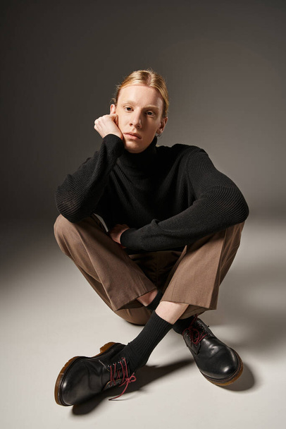 good looking young non binary model sitting on floor with crossed legs and hand near face, fashion - Photo, Image