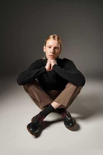 vertical shot of young non binary model in black turtleneck sitting on floor with crossed legs - Photo, Image