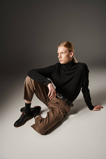 persona no binaria de buen aspecto en cuello alto negro y pantalones marrones sentados en el suelo, la moda - Foto, imagen