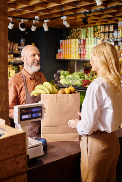 食料品店で新鮮なフルーツを購入する彼の顧客に笑顔のひげを持つ成熟したジョリーセラー - 写真・画像