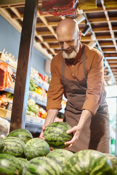 fröhlicher bärtiger älterer Verkäufer lächelt glücklich und pflückt frische Wassermelone im Supermarkt - Foto, Bild