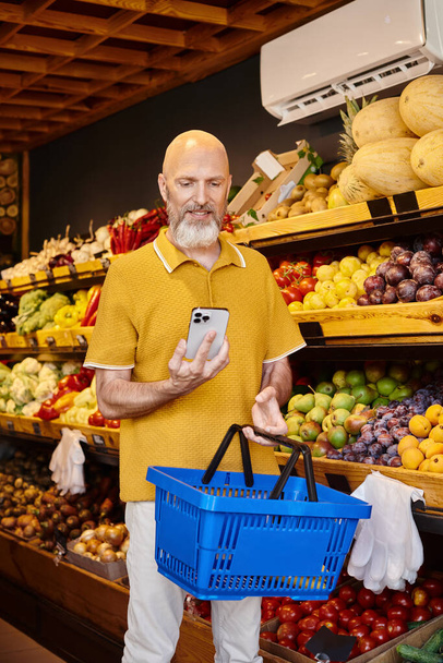 веселий бородатий клієнт тримає кошик для покупок і радісно посміхається по телефону в продуктовому магазині - Фото, зображення