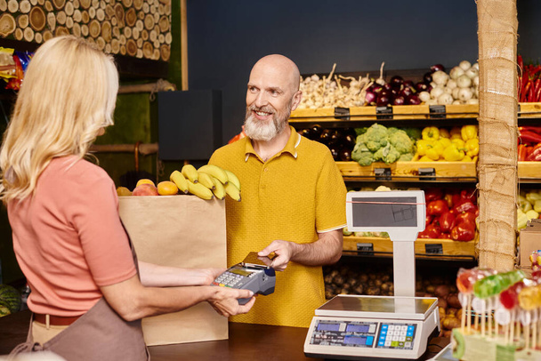 focus op vrolijke klant betalen met zijn telefoon voor vers fruit en glimlachen naar wazig verkoper - Foto, afbeelding