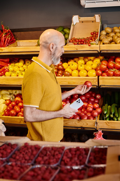 grijs bebaarde volwassen klant houdt mobiele telefoon en op zoek naar verse sappige tomaat in de supermarkt - Foto, afbeelding