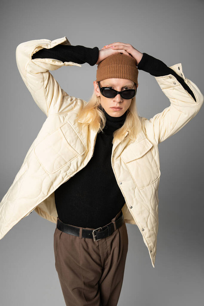 молодой небинарный человек в стильной зимней куртке позирует с руками на голове и смотрит в камеру - Фото, изображение