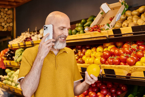 fröhlicher, graubärtiger Mann, der mit dem Handy telefoniert, während er sich im Supermarkt frisches Obst aussucht - Foto, Bild
