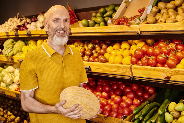 homme barbu gris joyeux en tenue décontractée posant avec un énorme melon dans les mains et souriant à la caméra - Photo, image