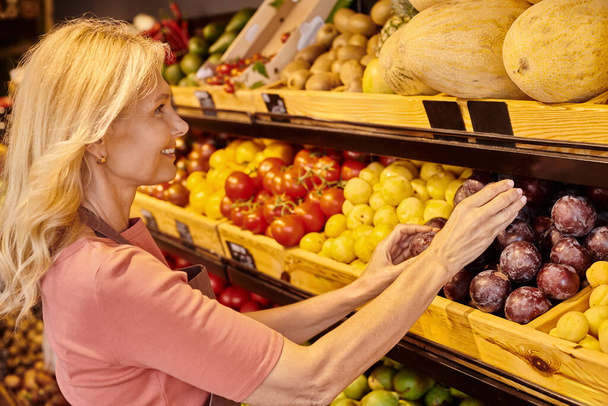 alegre madura vendedora poner fresco vibrante ciruelas en puesto con otras frutas en la tienda de comestibles - Foto, Imagen