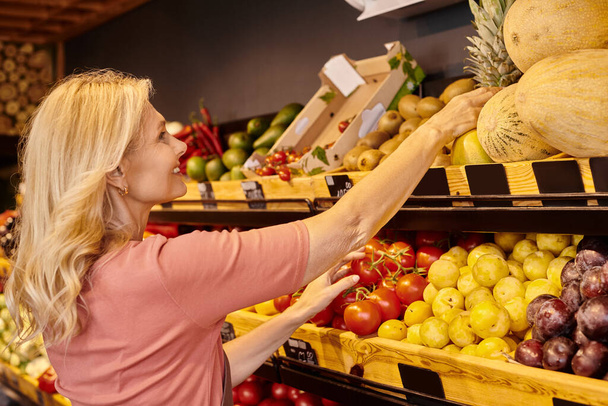 зрелый веселый продавец улыбается во время работы рядом с продуктовым ларьком со свежими фруктами и овощами - Фото, изображение