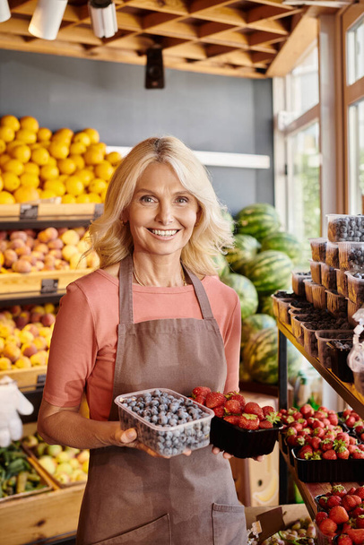 freudige reife Verkäuferin mit frischen, lebendigen Erdbeeren und Blaubeeren und lächelt in die Kamera - Foto, Bild