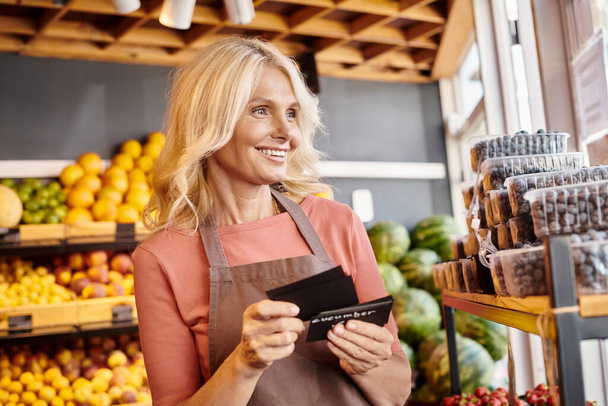 alegre vendedor maduro sosteniendo etiquetas de precio en las manos sonriendo felizmente y mirando hacia otro lado en la tienda de comestibles - Foto, imagen