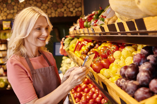 joyeux attrayant vendeur mature prenant des photos sur le téléphone de fruits et légumes frais et vibrants - Photo, image