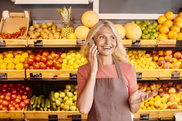 alegre vendedor atractivo sonriendo y hablando por teléfono con frutas y verduras en el telón de fondo - Foto, Imagen