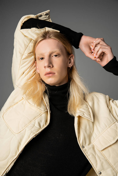 houkutteleva androgyyni henkilö talvella tyylikäs takki poseeraa harmaalla taustalla kädet pään yläpuolella - Valokuva, kuva