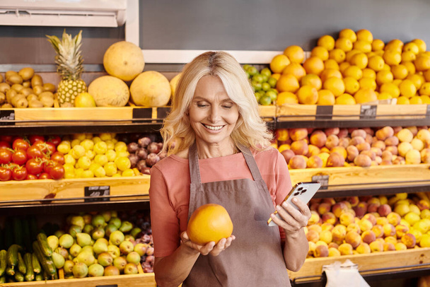 attraktive lustige Verkäufer halten Handy und frische nahrhafte riesige Orange und lächeln glücklich - Foto, Bild