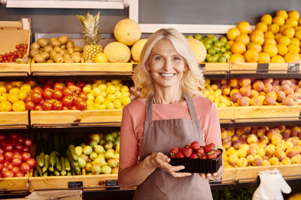 feliz madura vendedora posando con deliciosas fresas en las manos y sonriendo alegremente a la cámara - Foto, imagen