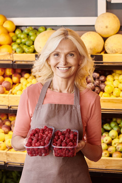 vertical shot of cheerful seller posing with packs of raspberries in hands and smiling at camera - Photo, Image