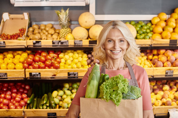 alegre vendedor maduro con bolsa de papel llena de verduras en las manos sonriendo a la cámara en la tienda de comestibles - Foto, Imagen