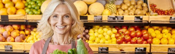 joyful mature seller with greens smiling at camera with fruits and vegetables on backdrop, banner - Photo, Image