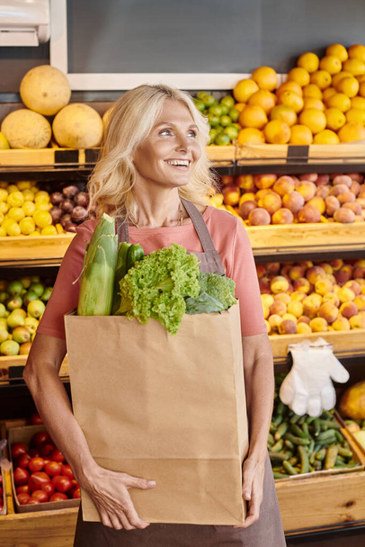 Reife, gut gelaunte Verkäuferin posiert mit Papiertüte voller Grünzeug und schaut weg vom Supermarkt - Foto, Bild
