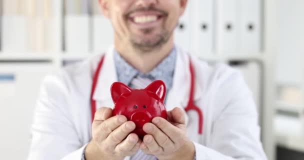 Genç gülümseyen doktor kırmızı domuz kumbarasını tutuyor. Sağlık sigortası kavramı - Video, Çekim