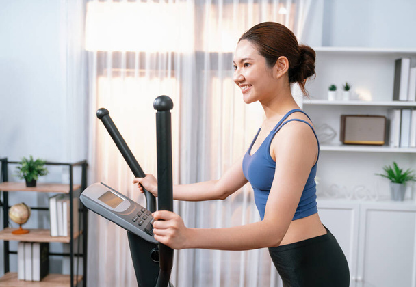 Femme asiatique athlétique énergique et forte qui court sur une machine elliptique à la maison. Poursuite de la forme physique et l'engagement à un mode de vie sain avec l'entraînement à domicile et la formation. Vigoureux - Photo, image