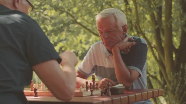 Waist up of two mature grey haired Caucasian men sitting at table in park on sunny day playing chess - Footage, Video