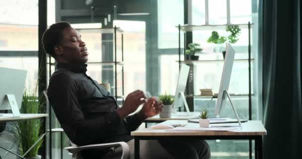 Een lachende zwarte zakenman zit achter een bureau in het kantoor. Zijn vrolijke houding en positieve houding zijn duidelijk zichtbaar in deze scène van professioneel geluk en vertrouwen. - Video