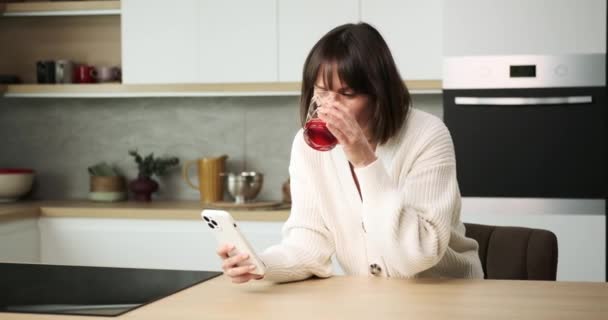 Una mujer caucásica está absorta en los mensajes de texto en su teléfono, mientras que casualmente sorbiendo jugo de un vaso. Su multitarea relajada refleja su capacidad para mantenerse conectado mientras disfruta de una bebida refrescante. - Metraje, vídeo
